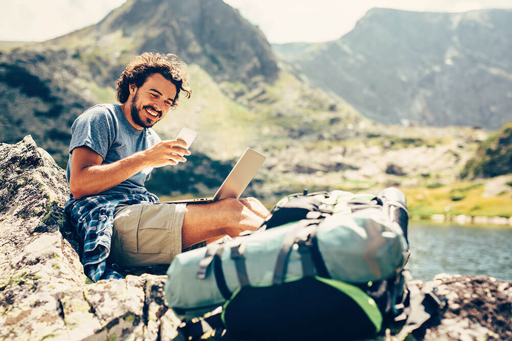 A man smiles as he works as a digital nomad from a beautiful location next to mountains and water